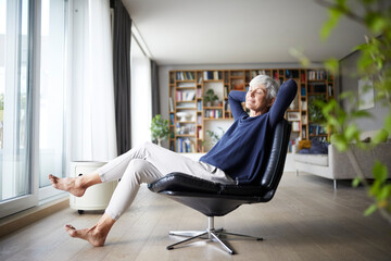 Contemplating senior woman with hands behind head sitting on chair at home