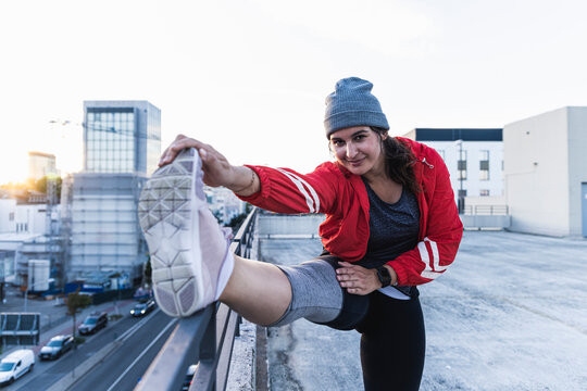 Young woman stretching leg on railing while standing on terrace against clear sky