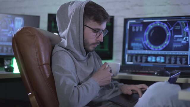 Side view of confident Caucasian programmer typing on laptop keyboard and drinking coffee at night. Portrait of concentrated young man hacking online web site. IT specialist overworking indoors.