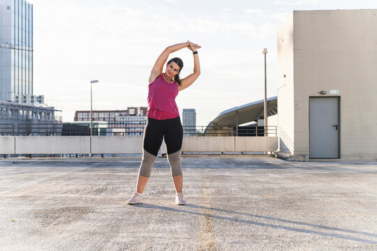 Young woman with arms raised exercising while standing on terrace against sky