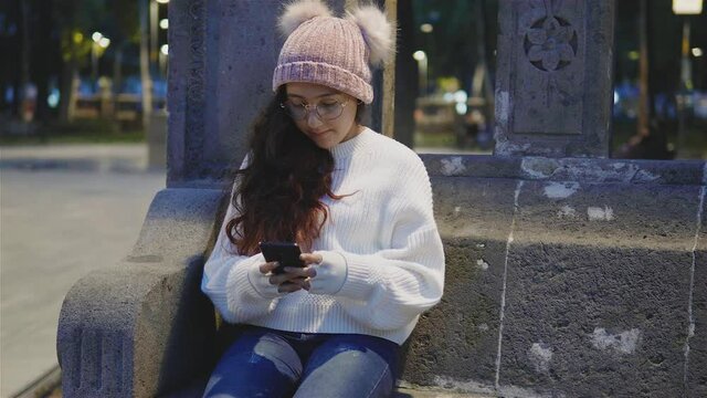 niña bonita,alegre vestida con ropa de invierno sentada tomándose fotos y mandando mensajes en el celular al aire libre