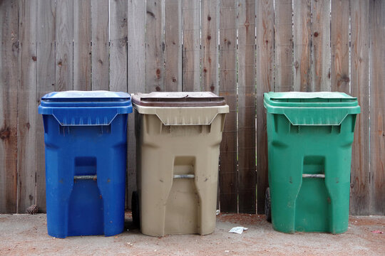 Three waste bins for recycling, garbage disposal, and gardening waste, side by side in front of a large wooden wall