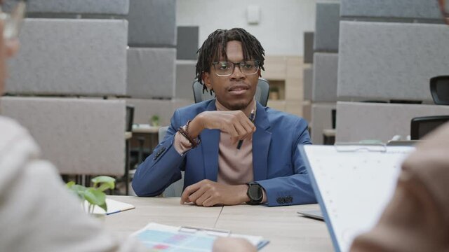 Rear view of young African businessman wearing blue suit sitting at desk in modern office in front of two diverse employees and giving task to them