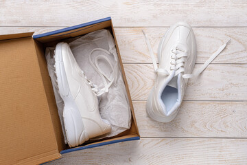 One white chunky sole sneakers in a brown cardboard box, the other on the white wood floor. Open box with new comfortable shoes for active lifestyle, fitness and sports.