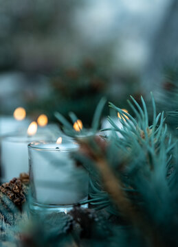 five lit white votive candles burning flames amid green pine branches and decorative pine cones
