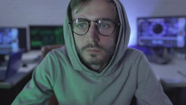 Close-up of concentrated man in eyeglasses looking at camera with multiple computers at the background. Portrait of absorbed young programmer or hacker in hood. Computer screen POV.