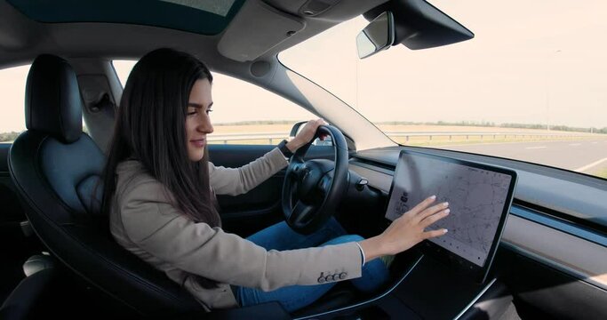 Beautiful Caucasian young woman sitting in electric vehicle and typing on big touch screen. Cheerful pretty female driver tapping on-board computer in car and searching route on GPS navigation map
