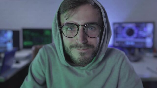Computer POV, happy young man in eyeglasses smiling and looking at USB plug on cable. Satisfied computer programmer or hacker at the background of multiple computers in dark room.