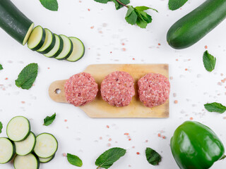 set of food.
Three raw cutlets on a wooden board in the middle, and along the edges zucchini, pepper, salt and herbs on a white background, close-up top view.