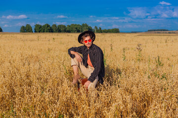 Naklejka na ściany i meble Tall handsome man with black hat and pink sunglasses sitting on a brown at golden oat field.