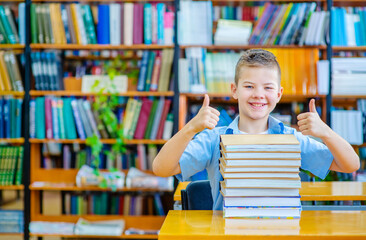 A boy in the library sits against the background of shelves with books and holds a stack of books in his desk with a smile showing a thumb up. Back to school