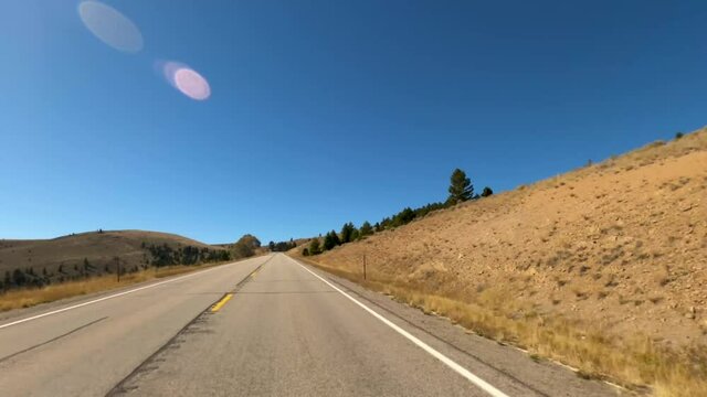 Driving a two-lane weathered asphalt road through the rolling hills of southwest Montana on Highway 287 passing dry autumn grass and scattered pines with sun flare-Drive Plate-POV