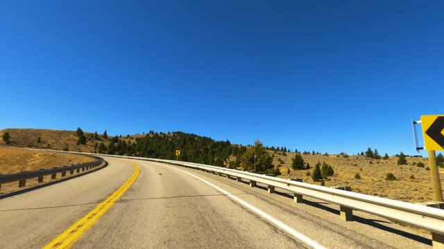 Driving Highway 287 Curving weathered two-lane asphalt highway in southwest Montana passing dry amber autumn grass and green pine trees under a clear blue sky-Drive Plate-POV