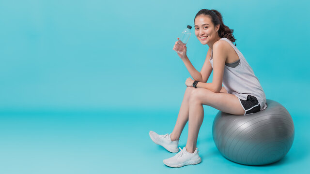 Asian beautiful happy woman holding water bottle and sitting on fit ball after exercise isolated on blue colour background.Concept of slim and healthy girl workout.