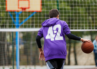 Cute young boy plays basketball on street playground. Teenager in purple hoodie with orange basketball ball outside. Hobby, active lifestyle, sport activity for kids.