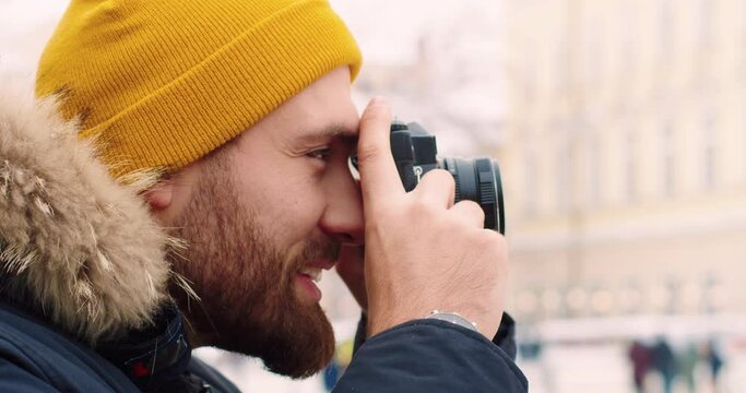 Close up of young bearded tourist in winter clothes making photo of city on camera. Winter travel.