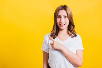 Fototapeta premium Asian Thai happy portrait beautiful cute young woman standing wear white t-shirt pointing finger away side looking to away side, studio shot isolated on yellow background with copy space