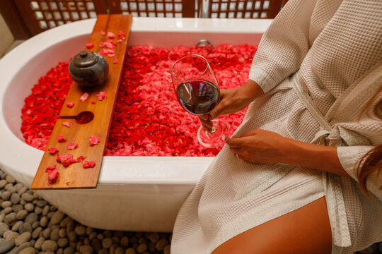 Cropped image beautiful woman in bathrobe sit near bath tub with rose petals drinking wine. Hands with galss of red wine close up