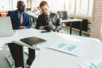 Mature afroamerican businessman to discuss information with a younger colleague. People working and communicating while sitting at the office desk together with colleagues sitting.