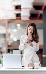 Portrait of young asian woman using smartphone while standing at her office desk in modern office.