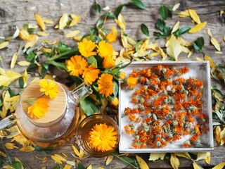 Medicinal autumn background. Herbal healthy marigold tea with a teapot, dried and fresh flowers on a wooden background with autumn leaves.