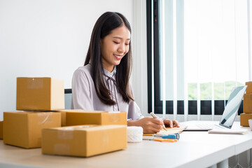 Young Asian woman business owner with many parcel boxes on the table happy online sales job, use your laptop, get an order from customers, take notes, and make arrangements for delivery by post