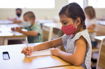 Portrait of diligent schoolgirl wearing protective face mask sitting in class writing exercise, new normal education during pandemic