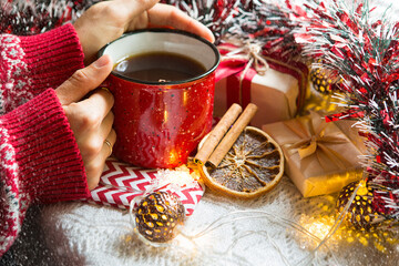 A woman's hand in a warm sweater holds a red mug with a hot drink on a table with Christmas...