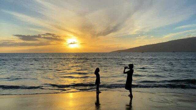 Silhouette of woman and young girl in dresses having fun taking photos in the water at a tropical beach at sunset. Calm ocean fills the frame, and a distant island is seen on the horizon with the sun.
