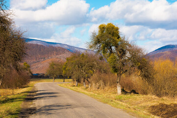 old road through countryside in early spring. leafless trees along the way. snow capped mountain in the distance. sunny weather with clouds on the sky