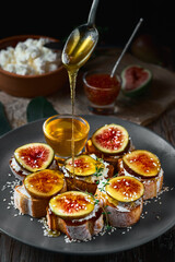 Honey dripping from a spoon onto fig sandwiches on toasted goat cheese baguette on a plate with honey, sesame seeds and walnuts. Fresh toast with figs and garnished with thyme leaves. Close-up