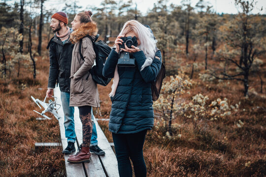 Beautiful blond girl with camera shots autumn relaxed forest staying on wooden path behind her friends.