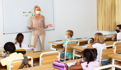 Young female teacher in protective face mask giving lesson to children in elementary school. New life reality during coronavirus pandemic