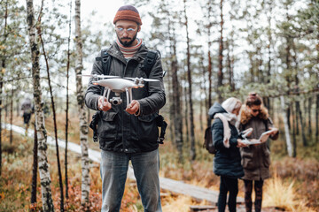 Lovely and relaxed autumn forest and bearded guy he holds quadcopter and two girls they stay behind.