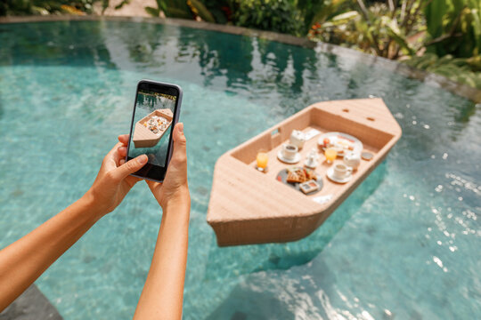 Woman taking picture on phone of floating breakfast boat shaped tray in swimming pool