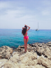 
The girl is photographed with her back, standing on the stones in a swimsuit. In the background a yacht, the sea.