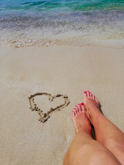 
A heart is drawn on the sand. The legs of the girl, the sea, summer are visible.