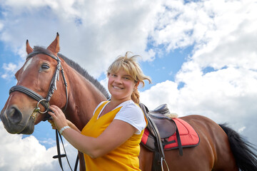 Obraz premium Girl in yellow dress with a horse on a green field and a blue sky with white clouds on the background