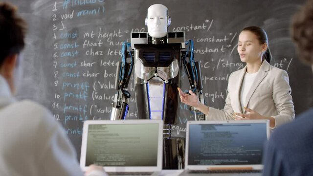 Medium shot of young mixed race female pupil making report at blackboard next to electronic robot to classmates working on laptops at bright equipped classroom