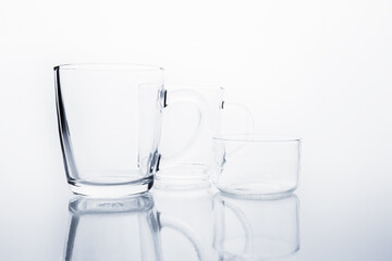 Two empty transparent glass mugs and saucer rosette on reflective surface on white background. Black and white transparent glass still life.
