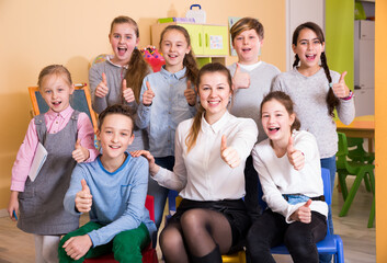 Cheerful group of pupils with female teacher posing together in schoolroom, giving thumbs up