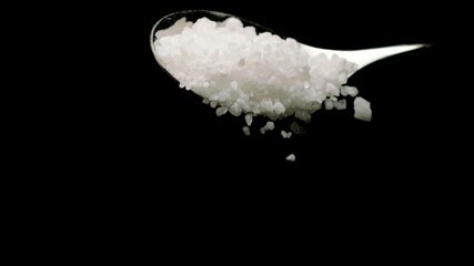 Full spoon of white sea salt. Coarse rock salt mineral grains falling from spoon in slow motion isolated on black background. Food, healthy eating, halite, nutrition, harm benefits of salt concept