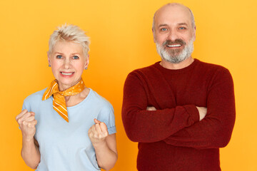 Displeased middle aged female with short dyed hair clenching fists being angry at her careless unfaithful bearded husband who crossing arms on his chest, smiling, having confident expression