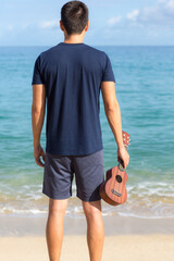 Young man holding an ukulele guitar standing on the beach looking at the ocean horizon.