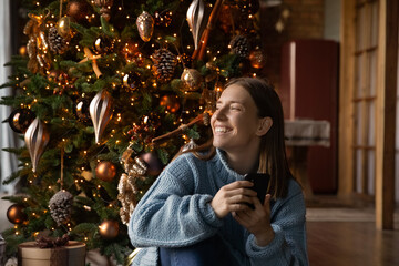 Happy dreamy millennial woman sit near Christmas tree use cellphone look in distance making wish....