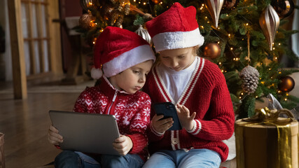 Wide banner panoramic view of little brother and sister kids have fun play on gadgets on winter holidays at home. Small boy and girl children look at screen enjoy online game use devices together.