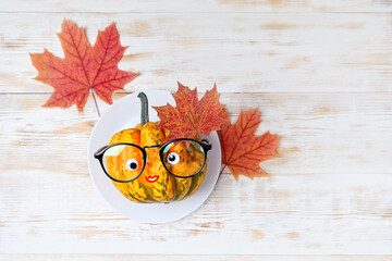 funny pumpkin in eyeglasses on plate with maple leaves, on white wooden table. fall harvest season,...