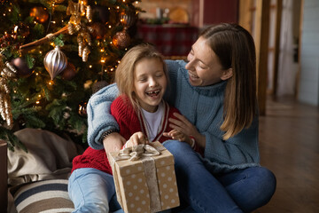Fototapeta na wymiar Happy loving young Caucasian mother greeting smiling small girl with Christmas present wrapped gift. Caring mom congratulate overjoyed little daughter with New Year, celebrate winter holidays at home.