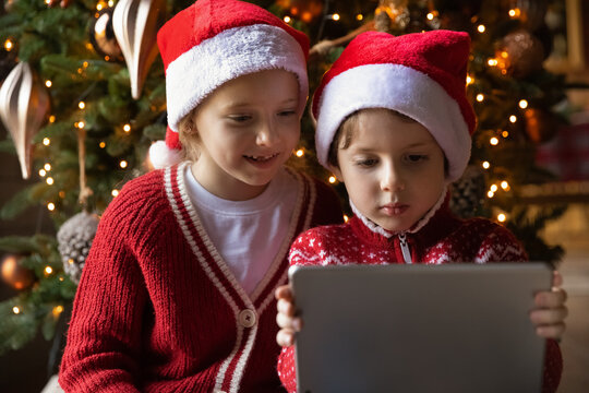 Close up of cute little boy and girl children in red Santa hats have fun watch video on tablet gadget at home. Smiling small brother and sister kids enjoy winter holidays play on new pad gift present.