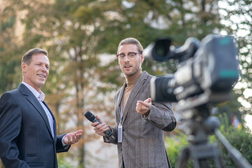 Fair-haired male journalist interviewing smiling mature male in front of camera